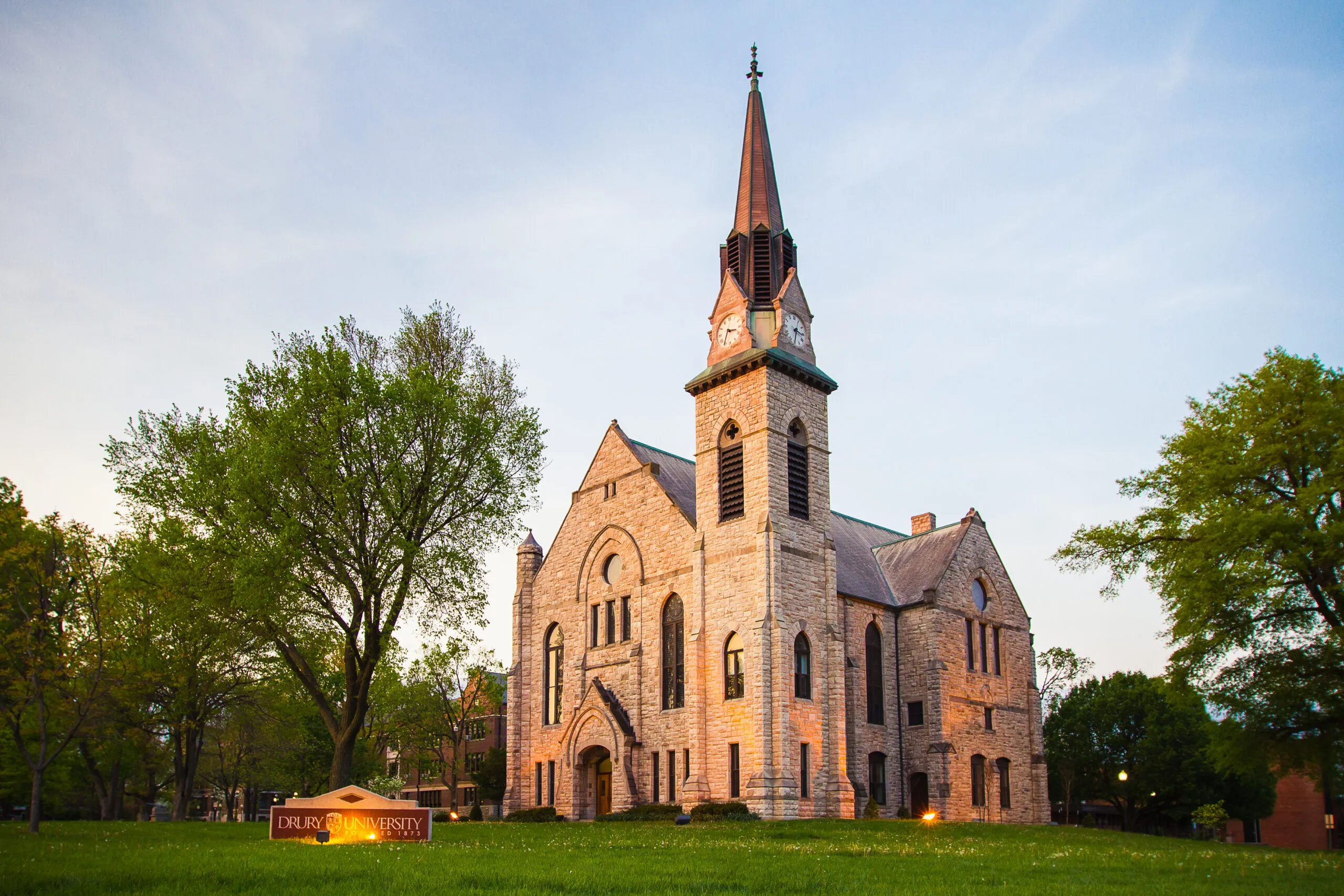 Stone Chapel at dusk, its limestone walls and clock tower lit against a blue sky, with the Drury University sign on the lawn in front.